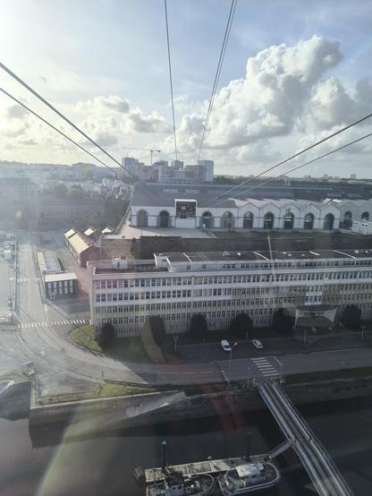 Blick aus der Seilbahn-Kabine: die Seile zeigen, dass die Mitte passiert wurde und es nun wieder runter geht zur Haltestelle. In der Bildmitte sind Gebäude, darunter der Fluss und auch ein Schiff, darüber der leicht bewölkte Himmel.