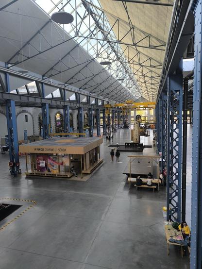 Das Bild zeigt eine große, offene Halle mit einem hohen, gläsernen Dach, das viel natürliches Licht hereinlässt. Die Halle ist mit einem robusten Stahlgerüst ausgestattet, das in blauen Farbtönen gestrichen ist. Im Zentrum der Halle befindet sich ein kleiner, quadratischer Ausstellungsraum. In der Halle sind mehrere Personen zu sehen, die entweder gehen oder sich auf Bänken ausruhen. Einige Bänke sind mit Tischen und Stühlen ausgestattet, auf denen Gegenstände wie Taschen und Kleidung liegen. Im Hintergrund sind weitere Ausstellungsstücke und eine große, gelbe Hebebühne zu erkennen. Die Halle hat einen industriellen Charakter, kombiniert mit modernen Elementen, und wirkt zugleich einladend und funktional.