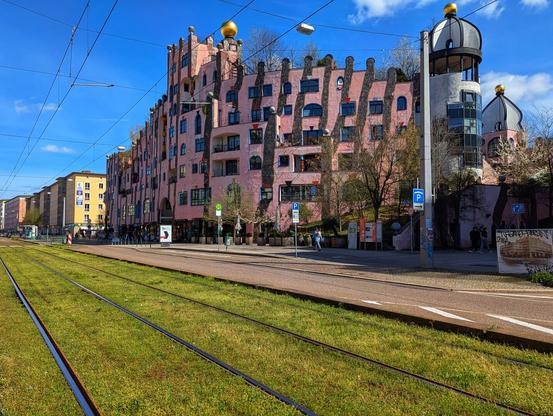 A vibrant, quirky pink building, the Grüne Zitadelle (Green Citadel), dominates the right side of the frame under a bright blue sky dotted with fluffy white clouds. Its undulating facade features numerous irregularly shaped windows and golden onion domes atop its towers. Trees with bare branches peek out from behind and on top of the building.
To the left, a more conventional yellow and grey apartment building stands in contrast. A grassy tramway track runs across the foreground, flanked by a grey pavement and a road with white lane markings. Several people are visible walking along the pavement and crossing the tram tracks. Parking signs are visible near the Grüne Zitadelle. Overhead tram wires crisscross the sky.