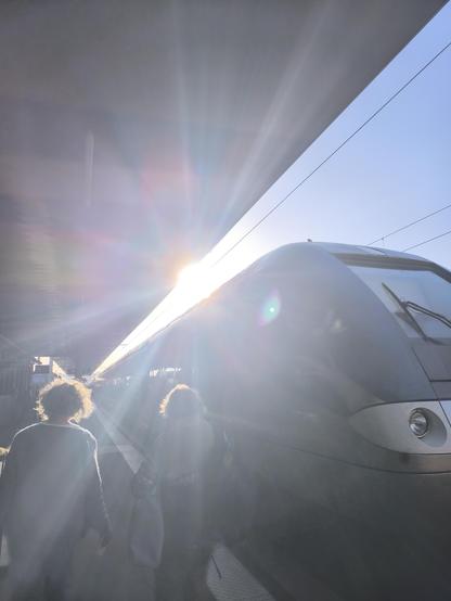 Die Sonne blendet direkt in die Kamera, aber man erkennt einen Zug, ein paar Menschen, die vor mir laufen und das Dach vom Bahnsteig. Vor allem aber scheint die Sonne!