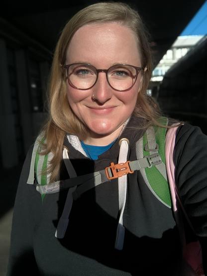Ein Portrait Foto von mir. Ich habe blonde lange Haare, eine Brille und ein Lächeln im Gesicht. Ich trage einen schwarzen Pulli und man erkennt die grünen Träger meines Rucksacks und die rosa Träger meiner Tasche. Die Sonne scheint mir ins Gesicht und der Schatten meines Handys in meiner Hand fällt mir auf die Brust.