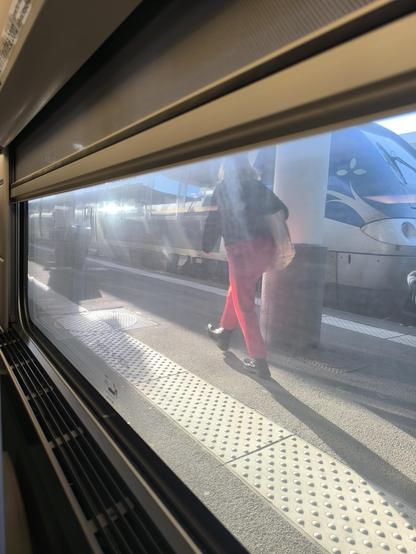 Blick aus dem TGV Fenster. Draußen ist der Bahnsteig, darauf läuft eine Person, dahinter steht ein anderer Zug.