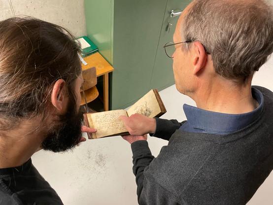 Zwei Personen schauen sich gemeinsam ein Stammbuch an. Man blickt über ihre Schultern. Die rechte Person zeigt auf eine Textstelle.