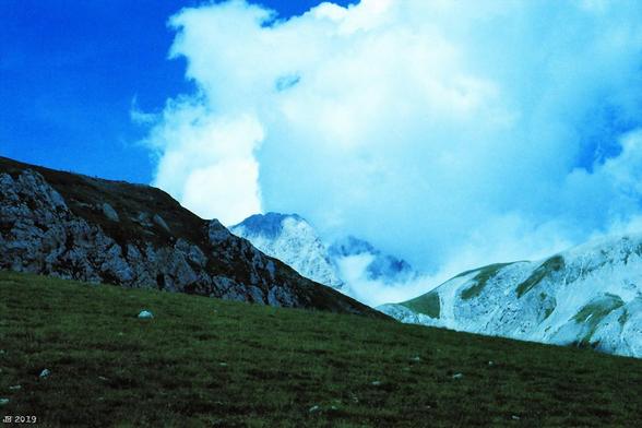 Blaustichiges Farbdiafoto, aufgenommen im Campo Imperatore in Abruzzo. In Bildmitte ein Teil des Gran Sasso, des höchsten Bergs in Mittelitalien. Im Vordergrund eine grüne Bergwiese mit vereinzelten weißen Steinen. Daran anschließend höhere Berge. Ab etwa Bildmitte bis rechts große weißblaue Wolken.