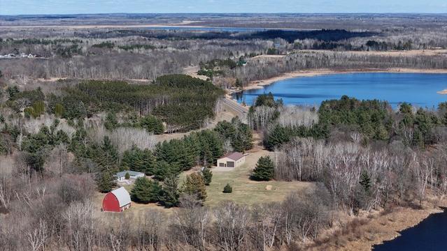 Aerial view of a small red barn in a spring landscape when most of the trees are still bare, except for groves of evergreens. There is a small lake in the background.