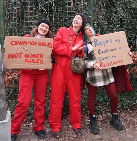 Smiling people in red outfits with cardboard signs saying "Cinnamon rolls NOT gender roles" and "Respect my existence or expect my resistance"