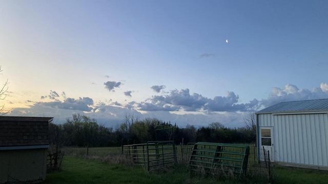 Looking across fields toward the southeast 15 minutes before sunrise. The sky’s color changes — left to right — from creamy yellow through several shades of blue. 

Dark blue clouds near the horizon. A white metal building has also a bluish hue.  

The crescent moon is visible in the blue sky.