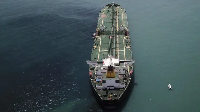 L'immagine mostra una petroliera di grandi dimensioni vista dall'alto, mentre naviga su un mare calmo. La nave è dipinta di verde scuro e bianco, e la sua struttura è chiaramente visibile. In lontananza, a destra, si vede una piccola imbarcazione. Il mare è di un colore blu-verde scuro.

Descrizione generata con AI.