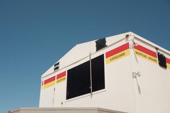 Low-angle, medium shot of a white, rectangular lifeguard station against a clear blue sky. The station has a pitched roof and a large, square window with a black opening. Horizontal bands of red and yellow run along the top edge of the station. Text within these bands reads "Lifeguard" and "Sauveteurs-Lifeguard." The roof has two small, square black vents. The station sits atop a light gray, concrete platform. The composition emphasizes the station's structure and signage.