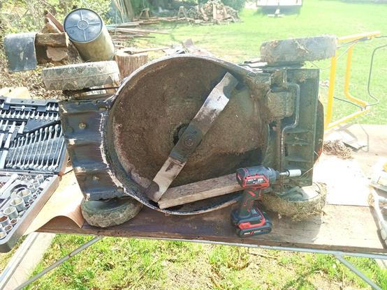 Underside of a lawn mower tilted on the side on an improvised workbench. The blade is exposed along with lots of dried grass choppings. A red and black impact driver with size 14 nut is standing in front of it.