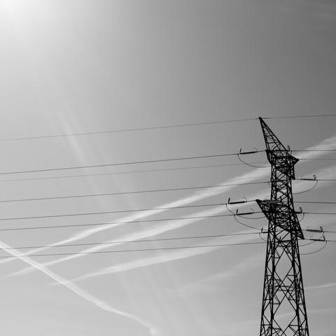A black and white photograph looking up at a tall power line tower on a bright, overcast day. Thin power lines stretch horizontally across the frame to and from the tower. In the background, faded contrails from airplanes crisscross the sky. The tower is heavily structured with a design of repeating geometric shapes, creating a latticework effect. The light, grayscale color palette gives the image a stark, industrial aesthetic.