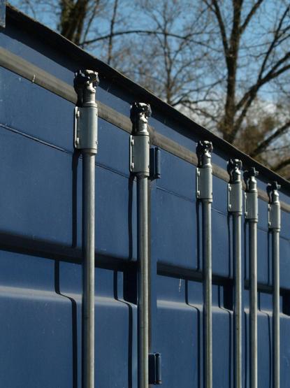 Low-angle, medium shot focusing on the side of a corrugated, navy blue shipping container. The container's vertical locking mechanism spans top to bottom with cylindrical, silver-colored metal poles and top mounted turning heads. The horizon line is visible in the upper third of the frame, with blurred tree branches and a blue sky in the background. Vertical lines of the poles are set against the horizontal lines of the container.