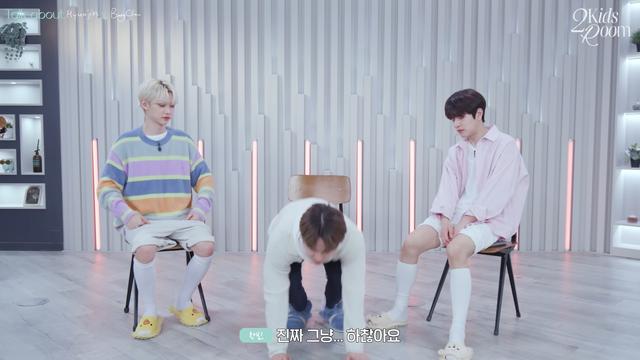 Felix and Seungmin sit on chairs either side of Changbin, who's left his chair to get on his hands and knees in front of them.