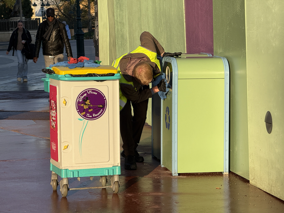 A Disney Village custodial cast member in a safety vest is cleaning a green recycling can, while a colorful trash can on wheels is nearby. The scene features pedestrians walking in the background, reflecting a lively environment.