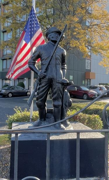 A bronze staue of Paul Rever in colonia garb and holding a rifle, on a pedestal, commemorating his historic ride that took him by this site. A U.S. flag stands behind  him.