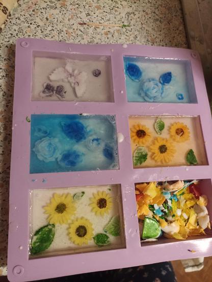 a tray of 6 silicone moulds for soaps where there are different soaps. in some there are sunflowers and leafs another two are with white and blue roses. the last one has butterflies in it.