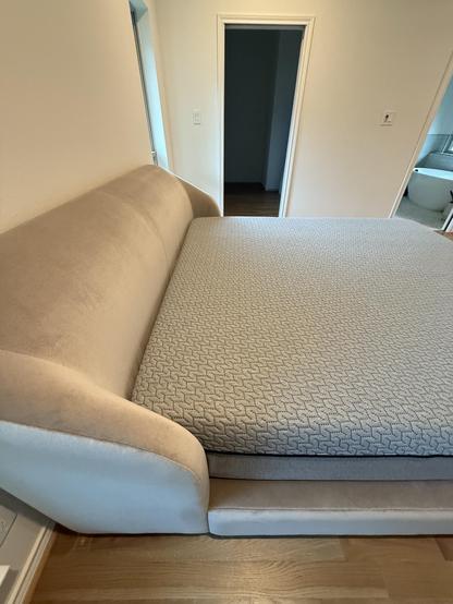A corner view of a modern, upholstered bed with a light beige headboard and a textured gray mattress. In the background, a doorway leads to another room, suggesting a spacious interior.