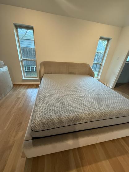 An empty bedroom featuring a modern bed with a light-colored upholstered frame and a patterned mattress. There are two angled windows allowing natural light, and a wooden floor adds warmth to the space. A glimpse of a fence and some outdoor greenery is visible through the
