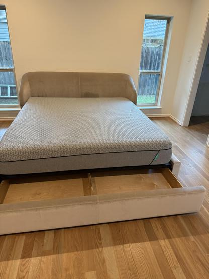 A contemporary bed frame with a plush headboard, featuring a queen-sized mattress partially inserted. The frame includes storage drawers beneath the mattress. The room has wooden flooring and two windows allowing natural light.