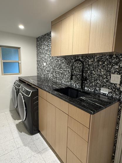 A contemporary laundry room featuring a countertop made of dark, marbled stone above a wooden cabinet. Two modern washing machines are visible, alongside a black sink. The backsplash has a textured, patterned design, and a small window allows natural light into the space