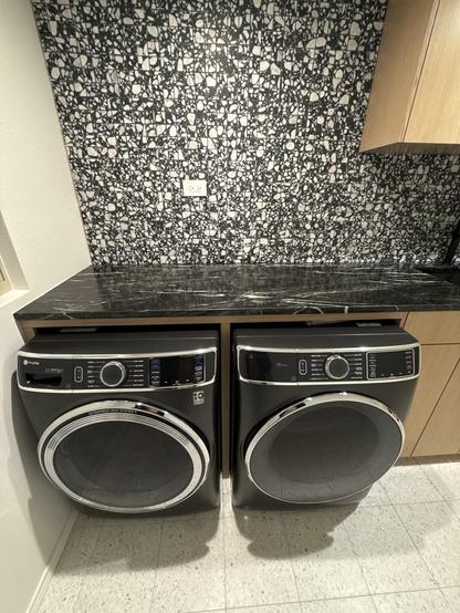 A modern laundry area featuring a stacked dryer and washing machine, both in sleek black design with chrome accents. They are placed beneath a dark marble countertop against a textured black-and-white wall. Natural light filters in from a nearby window.