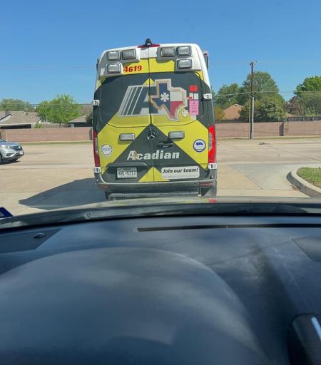 An Acadian ambulance is seen from behind, parked on a road. The vehicle features a bright yellow and white design with emergency lights on top. The view shows the interior of a vehicle, including the dashboard and steering wheel, positioned in front of the
