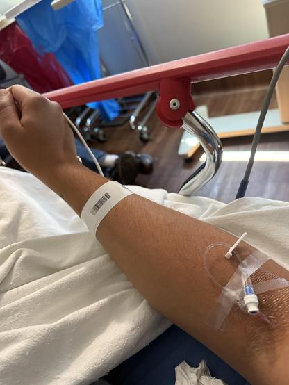 A close-up of an arm with a medical IV line attached, resting on a white blanket. A wristband is visible on the arm, and the background includes hospital materials and equipment.