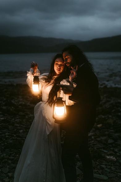 This atmospheric image, taken in Scotland during an elopement by Hamburg-based European elopement and wedding photographer Sturmsucht, captures a deeply intimate moment between a couple standing by what appears to be a Scottish loch or lake. 

The scene is set at dusk or nightfall, with dramatic dark clouds hanging low over distant mountains. The couple stands on a rocky shoreline, illuminated primarily by the warm glow of two vintage-style lanterns they're holding. The person on the left wears a flowing white wedding dress with billowy sleeves, suggesting they are the bride, while the person on the right is dressed in darker clothing. They stand very close together, their faces illuminated by the golden light of the lanterns, creating a striking contrast against the moody, dark background of water and mountains.

The soft lantern light creates an intimate, almost magical atmosphere against the otherwise dark, brooding Scottish landscape. The photograph beautifully captures the romance and adventure of an elopement in the Scottish wilderness, with the couple appearing as though they're in their own world, surrounded by nature's dramatic beauty.