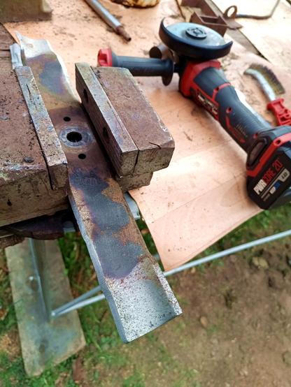 The mower blade, now detached from the rest of the mower and clamped into a big old drill press vise. A new cutting edge has been ground on. A red and black battery angle grinder with grinding disc is next to it.