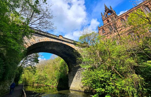 A tall arched stone bridge rising high above a river, with a distinctive red sandstone church alongside it.