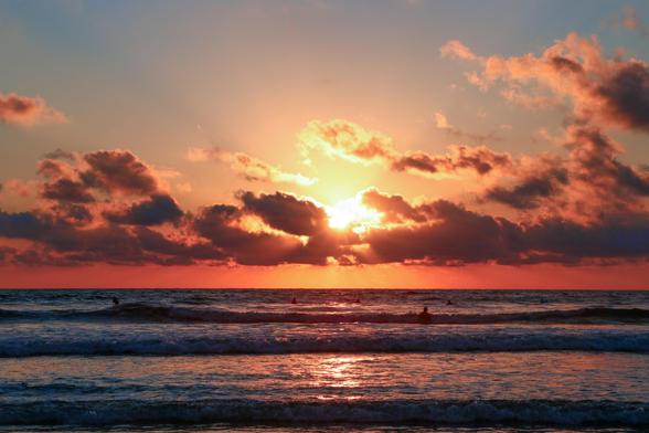 Sun sets through some clouds, over the ocean in Costa Rica. The sky is varied shades of orange, yellow, and blue. A lone swimmer is noticeable in the water in the distance.