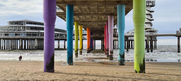 Photo of the pier on Scheveningen beach as seen from the beach under the walkway to the sea. The brown, black stained underside is on two parallel, brightly crayon colors colored rows of concrete pillars running straight into the sea, before forking to the left and right at a 90 degree angle. 

To the left a large three level, circular pavilion on several rows of grey, concrete pillars. To the right a smaller, one level pavilion with a white, round lookout tower also on a large group of pillars. A set of stairs snakes up around the tower to small platform with building with walkway around it.

To the left of the pier, barely visible, a person in a brown jacket and blue jeans crouches down at the foamy water's edge.