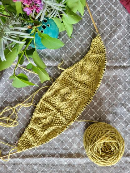 On a grey tablecloth with a light floral pattern lies a circular wooden knitting needle with the beginning of the back of a cardigan. It is textured with knit and purl stitches that create a diamond pattern. The yarn has a light dirty yellow colour. In the top left corner of the picture is a turquoise ball vase with some lavender and lilac.