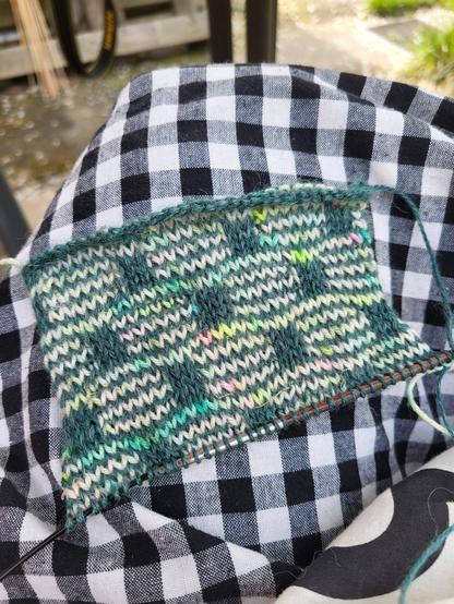 Medium close-up of the hem of my black and white gingham dress with a knitted swatch on it. The swatch is striped with two yarns: a teal one and a light one with neon speckles in green, yellow, pink and turquoise. The stripes are regularly interrupted by teal rectangles. The gauge is very small, needle size 3 mm.