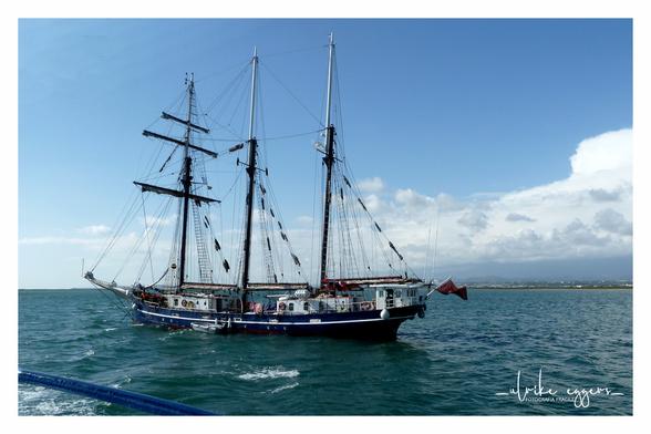 Ein altes Segelschiff mit blauem Rumpf und hohen Masten, aber ohne gesetzte Segel, auf dem offenen Meer an einem sonnigen Tag. Der Himmel ist hauptsächlich blau mit ein paar weißen Wolken, das Meer ist türkis. Im Hintergrund ist eine Küstenlinie mit Land zu sehen. Das Foto wurde von einem anderen Schiff aus aufgenommen, von dem im Vordergrund noch ein kleines Stück einer blauen Reling zu sehen ist.