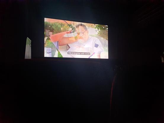 A dark cinema with a total shot of the big screen. The screen reveals a dissolve between two scenes featuring a film protagonist wearing glasses. The subtitle reads "Life is brighter with music."