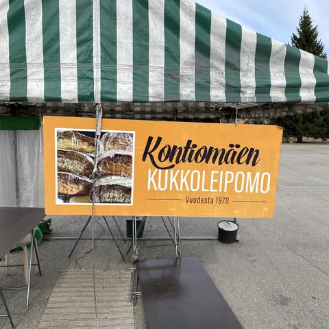 Torikoju: telttakatos, myyntipöytä, jakkara, matto maassa. Tori on tyhjä, päivä on päättynyt. Kojussa kyltti, jossa kuva kalakukkopinosta ja teksti "Kontiomäen kukkoleipomo. Vuodesta 1970".
