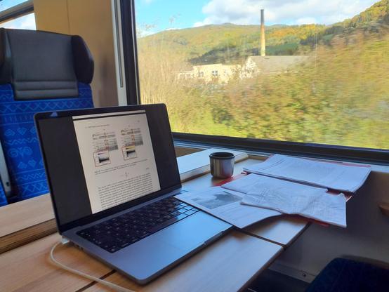 A picture from a train. There’s a laptop, various printed papers on the table, and a little cup, possibly with tea. Behind the train’s window is a blurred image of the landscape – green hills, blue sky and something resembling an old factory with a tall chimney.