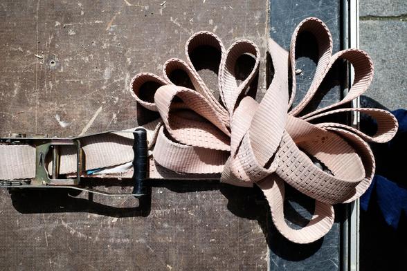 The image shows a beige ratchet strap neatly coiled into symmetrical loops on a rough wooden surface. The metal ratchet mechanism with a black plastic handle is visible on the left side, and sunlight casts sharp shadows across the scene, highlighting the strap’s woven texture.