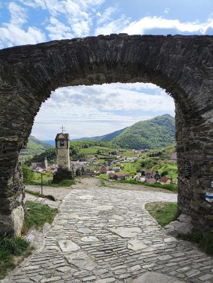 Eine alte Straße mit Pflastersteinen führt unter ein sehr altes Steintor durch. Dahinter sieht man ein Denkmal und dahinter eine Ortschaft im Tal.