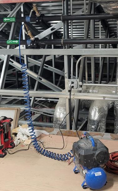 A small blue air compressor sitting on a wood floor. It's hooked to a piping system with a curly blue hose. In the background the galvanized steel roof trusses can be seen.