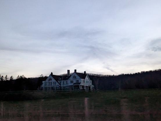 old, large abandoned house on top of a grassy hill surrounded by woods. very little sunlight left at twilight, no lights on in or around the house.