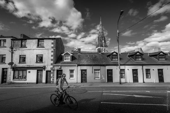 Black and white street photograph taken on Evergreen Street, Cork City, Ireland, showing a cyclist wearing sunglasses and a backpack riding past a row of terraced houses, with a church spire and dramatic clouds in the background; streetlamp and overhead wires visible, capturing an urban everyday moment.