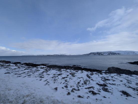 Snowy coastline, with cliffs in the background and a calm sea in between. Blue sky with very light clouds.