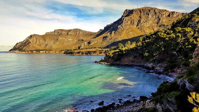 A scenic view of turquoise water meeting rocky cliffs and mountains under a partly cloudy sky.