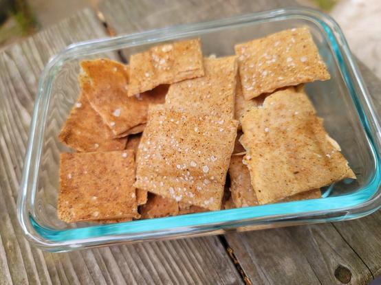 Sourdough Discard Crackers in a clear, rectangular glass container resting on a textured wooden surface. Inside, the roughly square crackers are nestled together. These baked crackers have a warm, light brown color with slightly darker edges in some places. Their surfaces appear somewhat rustic, with small speckles of sea salt and Cajun spices. The crackers vary slightly in size and shape, hinting at a homemade quality. The glass container has a subtle blue tint around its rim.