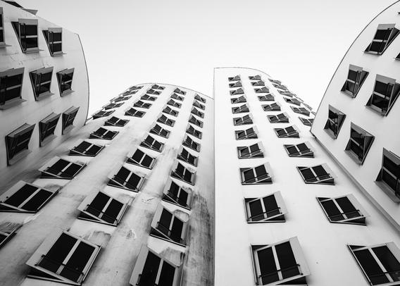 Black and white upward view of curving modern buildings with numerous identical windows, some slightly ajar, creating a dynamic rhythm. The architectural design features smooth, flowing façades that seem to lean inward, drawing the eye toward the pale sky above.