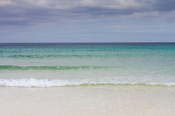 Picture shows a crystal clear blue/green sea with white sand a a blue sky.