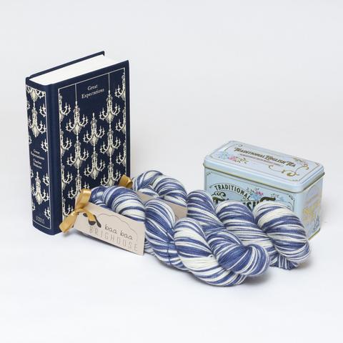 Picture shows two skeins of yarn in shades of blue and white positioned diagonally. To the left of the yarn, stood on its end, is a copy of the Penguin Clothbound Classic edition of Charles Dickens' Great Expectations. The book is navy blue with a repetitive cream chandelier patter on it. To the left of the yarn is a Victorian style pale blue tin of tea.