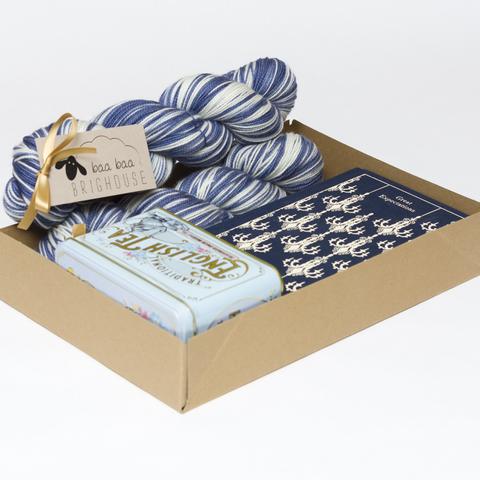 Picture shows an open A4 box containing two skeins of yarn in shades of blue and white. There is also a copy of the Penguin Clothbound Classic edition of Charles Dickens' Great Expectations. The book is navy blue with a repetitive cream chandelier pattern on it. There is also a pale blue Victorian style tin of tea.